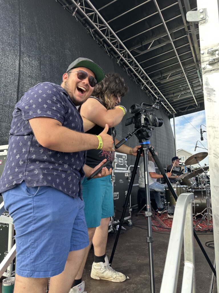 FTC student Fernando Gomez gives a thumbs up as he and John Maturo stand on the back of the Jambalaya stage to get video footage with a camera on a tripod 