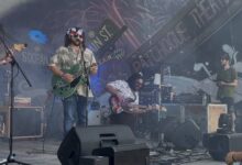 Three members of the band can be seen on a stage with a colofrul background during a performance